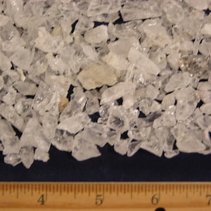 Puede incluir: Primer plano de un montón de pequeños cristales de cuarzo transparentes. Los cristales están esparcidos sobre una superficie azul y una regla de madera es visible en la parte inferior de la imagen. La regla está marcada con pulgadas del 3 al 7.