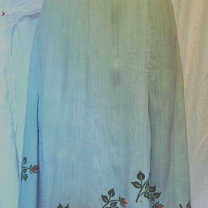 以下が含まれることがあります： 花柄のライトブルーのスカート。スカートはハイウエストで、わずかにAラインの形をしています。花柄は濃い緑の葉と赤いバラで構成されています。スカートは質感のある生地で作られています。