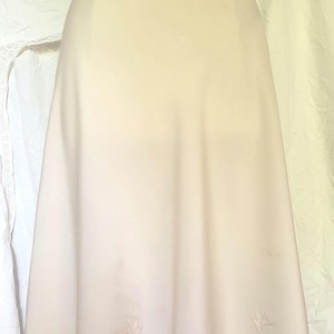 May include: A cream-colored vintage skirt with a scalloped hemline and embroidered leaf details. The skirt is displayed on a mannequin, showcasing its A-line shape and delicate design. The fabric appears to be a lightweight material.