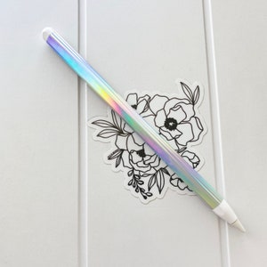 May include: A holographic stylus with a white tip and end cap, resting on a white surface. A black and white floral sticker is positioned behind the stylus. The stylus has a rainbow iridescent finish.