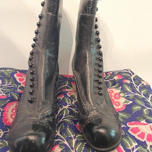 Puede incluir: Un par de botines negros vintage con cierres de botones. Los botines tienen una punta redondeada y un tacón pequeño. El cuero muestra signos de desgaste, lo que añade encanto antiguo. Los botines se exhiben sobre una tela estampada floral.