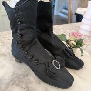 Peut inclure: Paire de bottines noires vintage. Elles présentent des boutons décoratifs, un nœud avec une boucle argentée et un petit talon. Les bottines sont en tissu souple. Hauteur approximative : 20 cm.