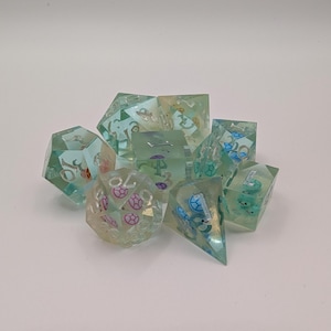 May include: A collection of translucent, multi-sided dice in shades of blue and green. The dice have embedded designs and numbers, with a few featuring pink and blue accents. The dice are arranged in a pile against a white background.