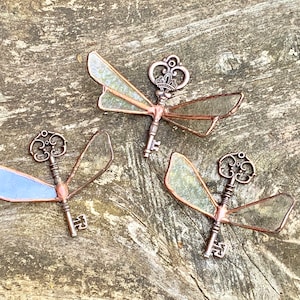 May include: Three decorative key-shaped dragonfly ornaments with translucent wings. The wings are made of stained glass and the keys are made of metal with a copper finish.