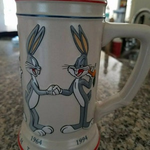 May include: A white ceramic Bugs Bunny stein with a handle, featuring Bugs Bunny characters shaking hands. The mug has red and blue accents, with the text "1964 THROUGH THE YEARS 1994".