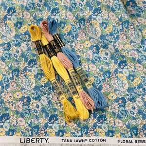 May include: A floral fabric with a blue, yellow, and pink pattern is shown with four spools of embroidery floss in yellow, pink, and blue. The fabric has the text "LIBERTY. TANA LAWN&trade; COTTON FLORAL REBELLION."