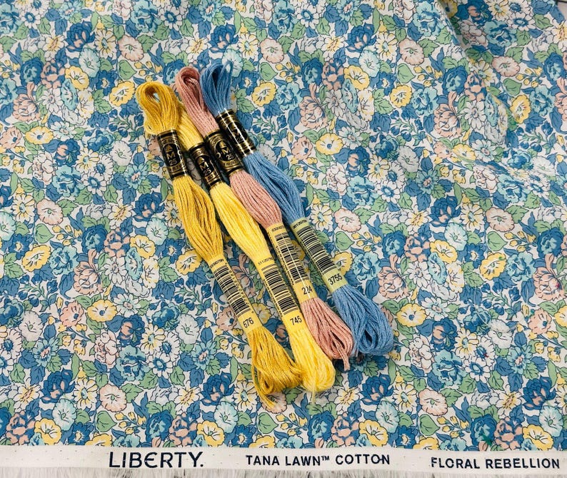 May include: A floral fabric with a blue, yellow, and pink pattern is shown with four spools of embroidery floss in yellow, pink, and blue. The fabric has the text "LIBERTY. TANA LAWN&trade; COTTON FLORAL REBELLION."