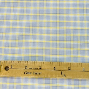 May include: A close-up of a blue and yellow plaid fabric with a wooden ruler measuring 1 1/8 yards. The ruler has the words "One Yard" printed on it.