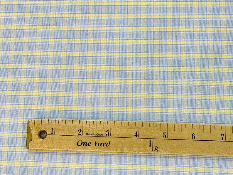 May include: A close-up of a blue and yellow plaid fabric with a wooden ruler measuring 1 1/8 yards. The ruler has the words "One Yard" printed on it.