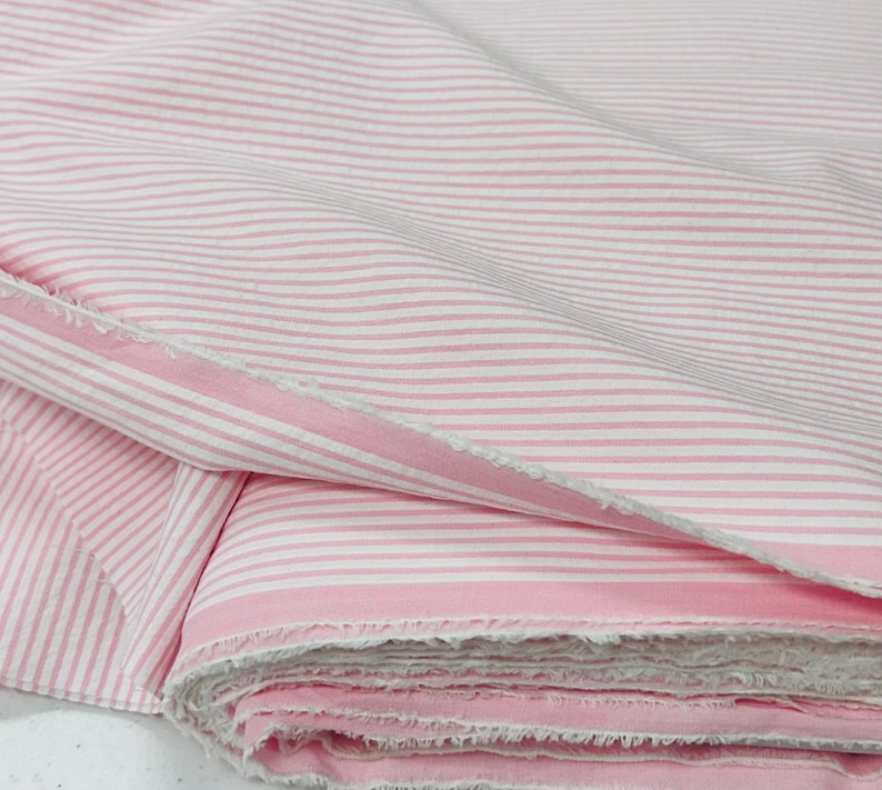 Puede incluir: Primer plano de una pila de tela a rayas rosas y blancas. La tela est&aacute; doblada, revelando los bordes sin rematar del material. Las rayas corren horizontalmente por la tela, creando un patr&oacute;n cl&aacute;sico.