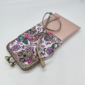 Puede incluir: Un estuche para gafas rosa y blanco con un diseño floral y de calaveras. El estuche tiene un marco de metal dorado y un cierre. Un par de gafas transparentes está dentro del estuche.