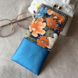 Può includere: Un astuccio rettangolare per occhiali con una sezione superiore in tessuto blu scuro con un motivo floreale di fiori arancioni, bianchi e azzurri. La sezione inferiore è in materiale blu metallizzato. Presenta una chiusura a scatto in bronzo.