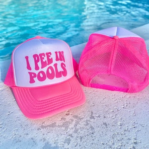 May include: Pink and white trucker hat with the text "I PEE IN POOLS" in pink letters. The hat is sitting on a white surface.