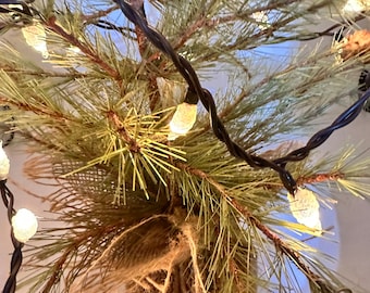 May include: A small artificial pine tree with burlap wrapped around the base. The tree is decorated with white string lights.