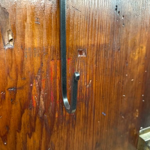 May include: A black metal hook is mounted on a wooden surface. The hook is bent into a J shape and is attached to the surface with a screw.