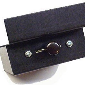 Puede incluir: Una pequeña caja negra con una perilla negra en la parte superior. La caja está abierta y se pueden ver dos tornillos plateados en la parte inferior.