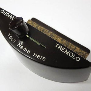 May include: A black and brown vintage tremolo pedal with a knob and the text "CHORA" and "TREMOLO". The pedal also has the text "Your Name Here" engraved on the front.