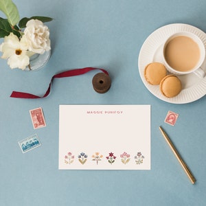 May include: A rectangular off-white stationery card featuring the name MAGGIE PURIFOY printed in red at the top and a row of six colourful floral illustrations along the bottom edge, accompanied by two vintage postage stamps and a gold pen.
