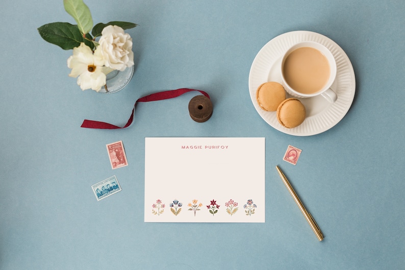 May include: A rectangular off-white stationery card featuring the name MAGGIE PURIFOY printed in red at the top and a row of six colourful floral illustrations along the bottom edge, accompanied by two vintage postage stamps and a gold pen.
