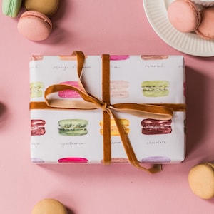 May include: A white gift box wrapped in paper with a pattern of colorful macarons. The box is tied with a brown velvet ribbon. The macarons are labeled with their flavors: pistachio, lemon, caramel, and chocolate.