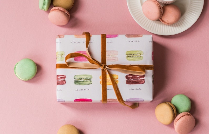 May include: A white gift box wrapped in paper with a pattern of colorful macarons. The box is tied with a brown velvet ribbon. The macarons are labeled with their flavors: pistachio, lemon, caramel, and chocolate.