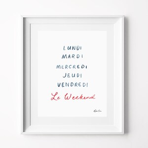 May include: A white framed wall art print featuring the French days of the week in dark blue handwritten text: Lundi, Mardi, Mercredi, Jeudi, Vendredi, followed by the phrase Le Weekend in red handwritten script, with a small signature at the bottom.