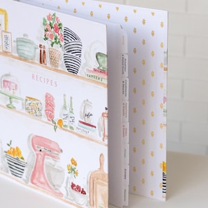 May include: A recipe binder with a watercolor design featuring kitchen shelves with various items. The binder has labeled dividers and a gold floral pattern on the inside cover. The word "RECIPES" is visible on the front.