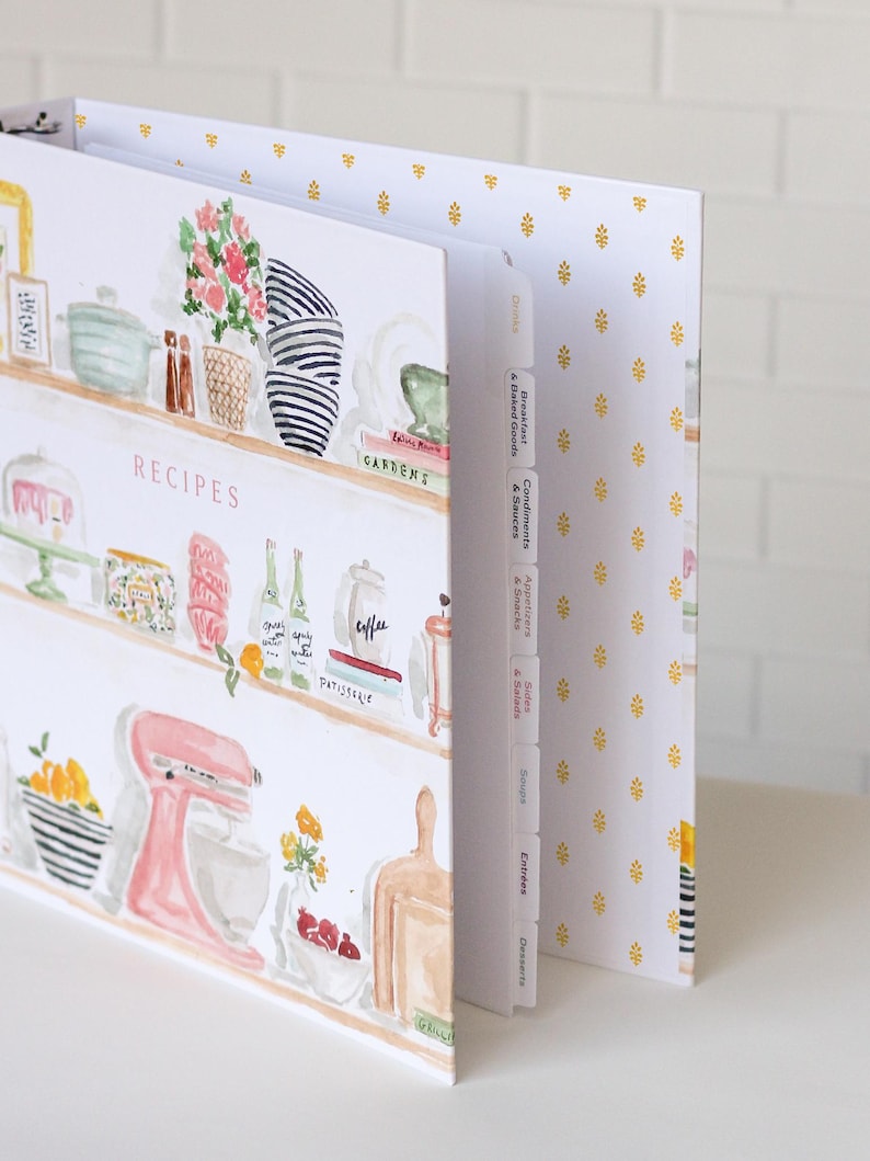 May include: A recipe binder with a watercolor design featuring kitchen shelves with various items. The binder has labeled dividers and a gold floral pattern on the inside cover. The word "RECIPES" is visible on the front.