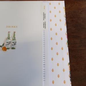 May include: A white binder with the word "DRINKS" and an illustration of two green bottles and an orange. The binder has tab dividers labeled "Drinks", "Breakfast & Baked Goods", and "Condiments & Sauces". The cover has a gold floral pattern.