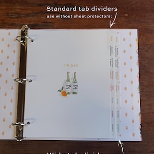May include: A white binder with gold accents and a floral pattern. The binder is open to a page with a watercolor illustration of two bottles and an orange. The page has tab dividers labeled "Drinks", "Breakfast & Baked Goods", "Condiments & Sauces", "Appetizers & Snacks", "Sides & Salads", "Soups", "Entrees", and "Desserts".