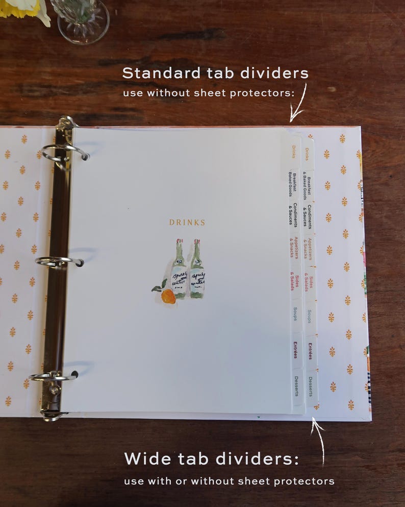 May include: A white binder with gold accents and a floral pattern. The binder is open to a page with a watercolor illustration of two bottles and an orange. The page has tab dividers labeled "Drinks", "Breakfast & Baked Goods", "Condiments & Sauces", "Appetizers & Snacks", "Sides & Salads", "Soups", "Entrees", and "Desserts".