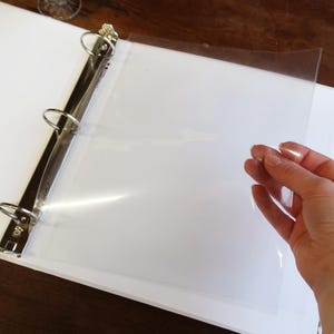 Peut inclure: Un classeur blanc avec des anneaux en métal argenté et une feuille de plastique transparente. Le classeur est ouvert et une main tient la feuille de plastique. L'arrière-plan est une surface en bois sombre.