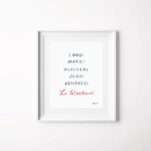 May include: A white framed wall art print featuring the French days of the week in dark blue text: Lundi, Mardi, Mercredi, Jeudi, Vendredi, followed by the phrase Le Weekend in red cursive script. A small signature is visible in the bottom right corner.