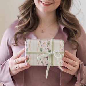 May include: A rectangular gift box wrapped in cream-colored paper featuring a vertical striped pattern and a floral motif with pink and green accents. The box is tied with a sage green velvet ribbon tied in a bow at the center of the front.