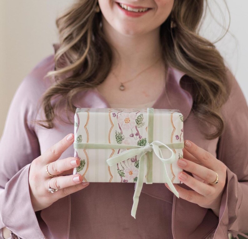 May include: A rectangular gift box wrapped in cream-colored paper featuring a vertical striped pattern and a floral motif with pink and green accents. The box is tied with a sage green velvet ribbon tied in a bow at the center of the front.