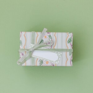 May include: A rectangular gift box wrapped in white paper featuring a pattern of vertical light gray stripes, orange wavy lines, and floral motifs. The box is tied with a light green velvet ribbon and includes a blank white rectangular gift tag.