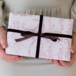 May include: A rectangular gift box wrapped in white paper featuring a repeating light pink illustration of dancers and trees, tied with a dark brown velvet ribbon arranged in a cross pattern and secured with a centered bow.