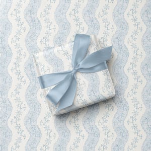 May include: A square gift box wrapped in white paper with a repeating pattern of light blue wavy vertical bands featuring small floral motifs. The box is tied with a smooth, light blue satin ribbon tied into a centered bow on the top surface.
