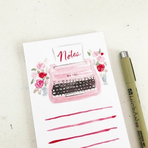 May include: A white notepad featuring a watercolour illustration of a pink Royal typewriter with the word Notes in red script above it. The typewriter is surrounded by pink and red flowers. Below the typewriter are four horizontal red lines.