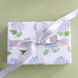 May include: A rectangular gift box wrapped in white paper featuring a repeating watercolor pattern of blue hydrangea flowers and green leaves. The package is tied with a shimmering silver satin ribbon arranged in a centered bow.