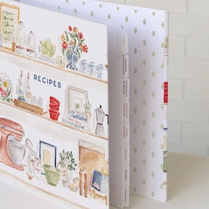 May include: A recipe binder featuring a watercolor illustration of kitchen shelves filled with cooking supplies, a stand mixer, and jars. The cover displays the word RECIPES in blue text. The binder includes white tabbed dividers labeled with categories.