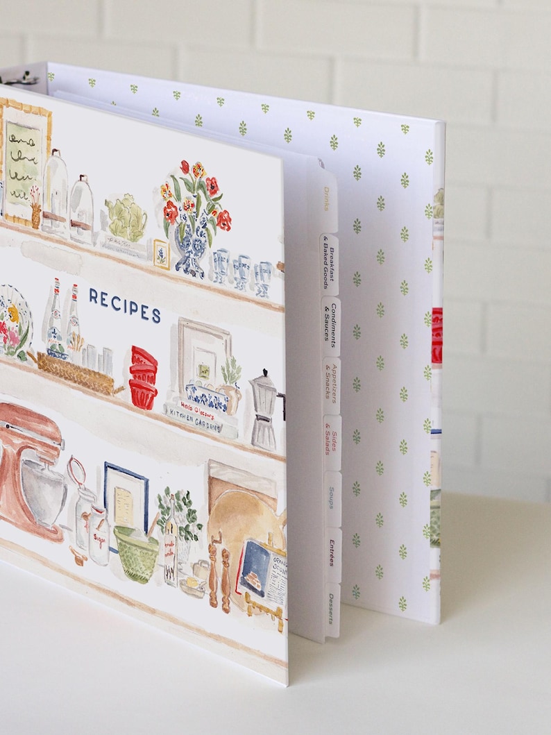 May include: A recipe binder featuring a watercolor illustration of kitchen shelves filled with cooking supplies, a stand mixer, and jars. The cover displays the word RECIPES in blue text. The binder includes white tabbed dividers labeled with categories.