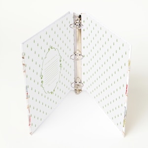 May include: An open three-ring binder with a white interior featuring a repeating green leaf pattern. The left side includes a decorative frame with lines for text that reads This book belongs to and Date. The metal ring mechanism is centered.