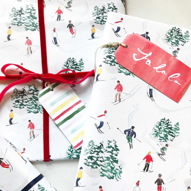 May include: White wrapping paper with a repeating pattern of skiers and pine trees. The paper is wrapped around a gift box with a red ribbon and a striped gift tag. A red gift tag with the text "Jo Jo" is attached to the paper.