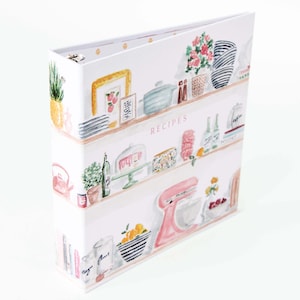 May include: A white recipe binder with a watercolor design of kitchen shelves. The shelves are filled with various kitchen items, including plants, framed art, and a pink stand mixer. The word "RECIPES" is printed on the front.