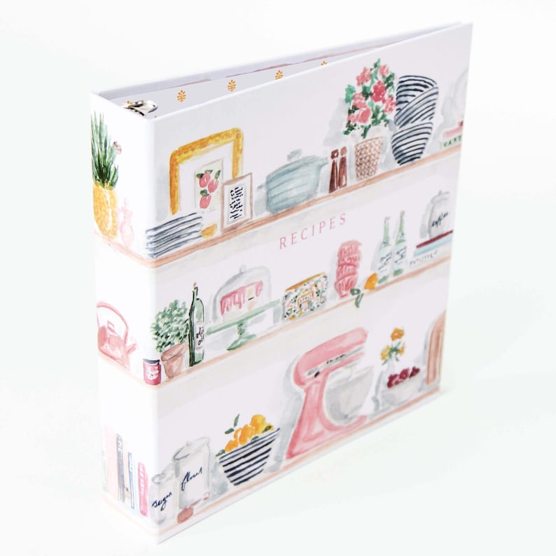 May include: A white recipe binder with a watercolor design of kitchen shelves. The shelves are filled with various kitchen items, including plants, framed art, and a pink stand mixer. The word "RECIPES" is printed on the front.