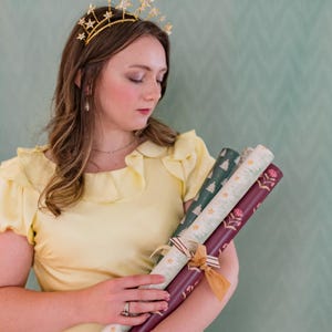 May include: A woman wearing a gold star crown and a yellow dress holds three rolls of patterned wrapping paper. The wrapping paper features designs of stars, Christmas trees, and floral patterns. The rolls are tied with ribbons.