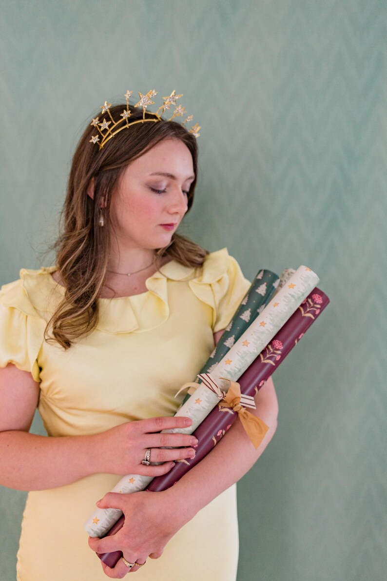 May include: A woman wearing a gold star crown and a yellow dress holds three rolls of patterned wrapping paper. The wrapping paper features designs of stars, Christmas trees, and floral patterns. The rolls are tied with ribbons.