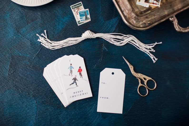 May include: A set of white gift tags with a watercolor illustration of people ice skating. The tags say "Merry Christmas" and have a space for "To" and "From".