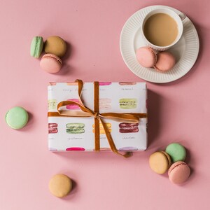 May include: A gift box wrapped in white paper with a brown ribbon and a pattern of colorful macarons. The box is surrounded by macarons in various colors, including pink, green, and brown. A cup of coffee and a saucer are in the upper right corner of the image.